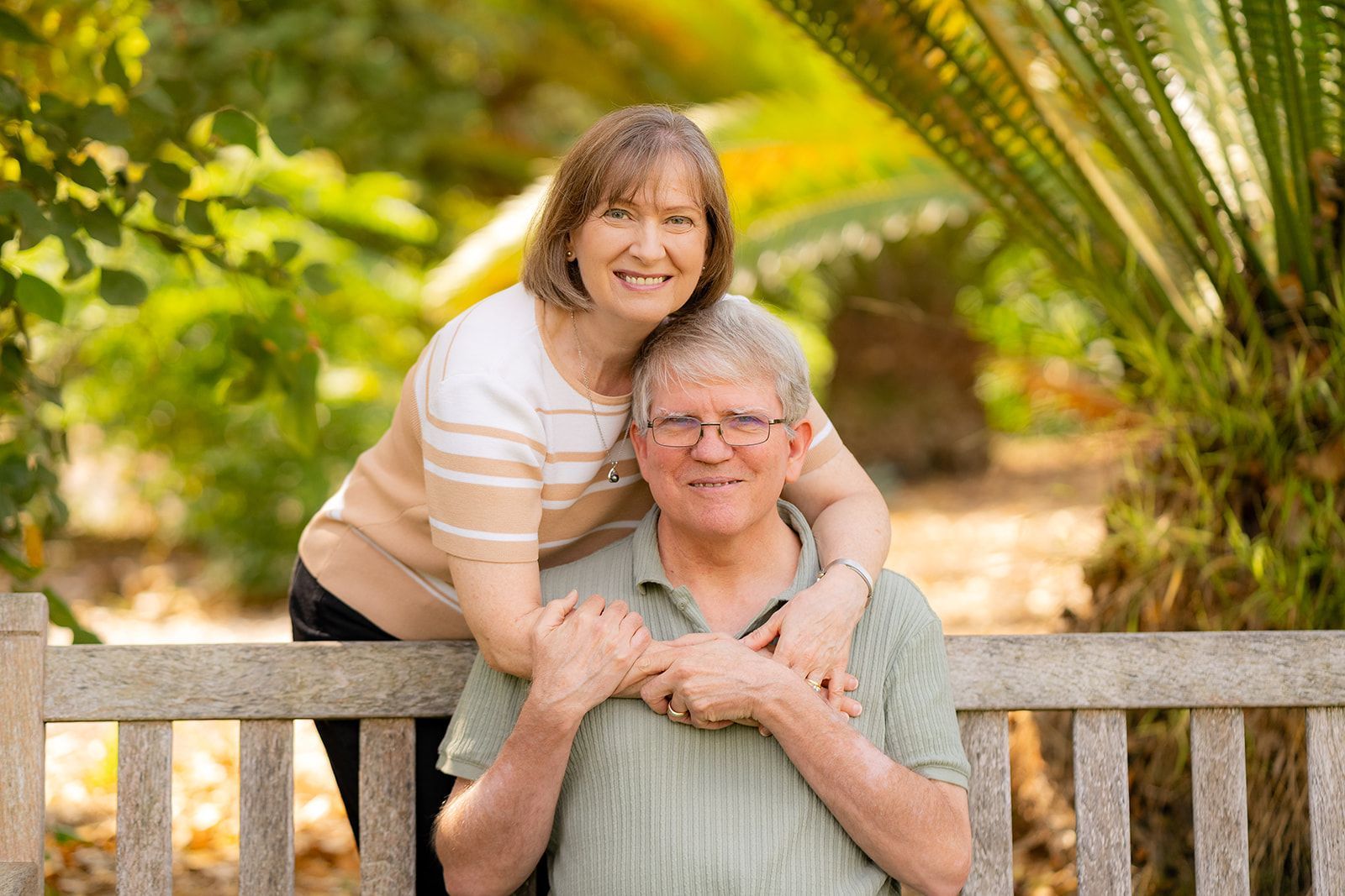 Fullerton Arboretum Family Portrait Woman embraces man sitting on a wooden bench outdoors; both smiling, greenery in background at Fullerton Arboretum