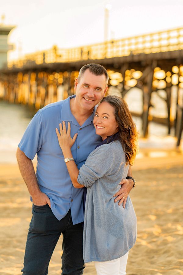 A man and a woman are posing for a picture on the beach in front of a pier during the sunset. 