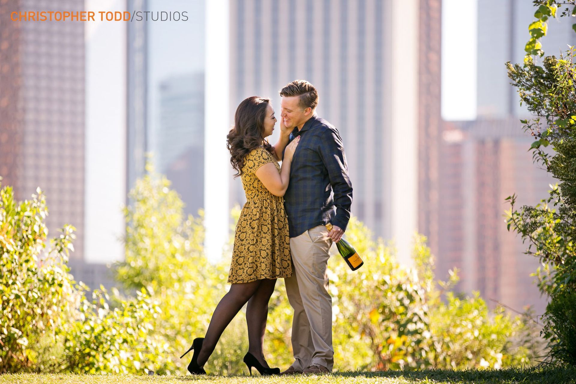 A man and woman are kissing in a park with the downtown Los Angeles skyline in the background.