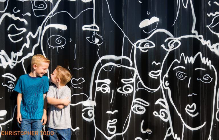 Costa Mesa Portrait Location Two boys are standing in front of a wall with drawings of faces on it.