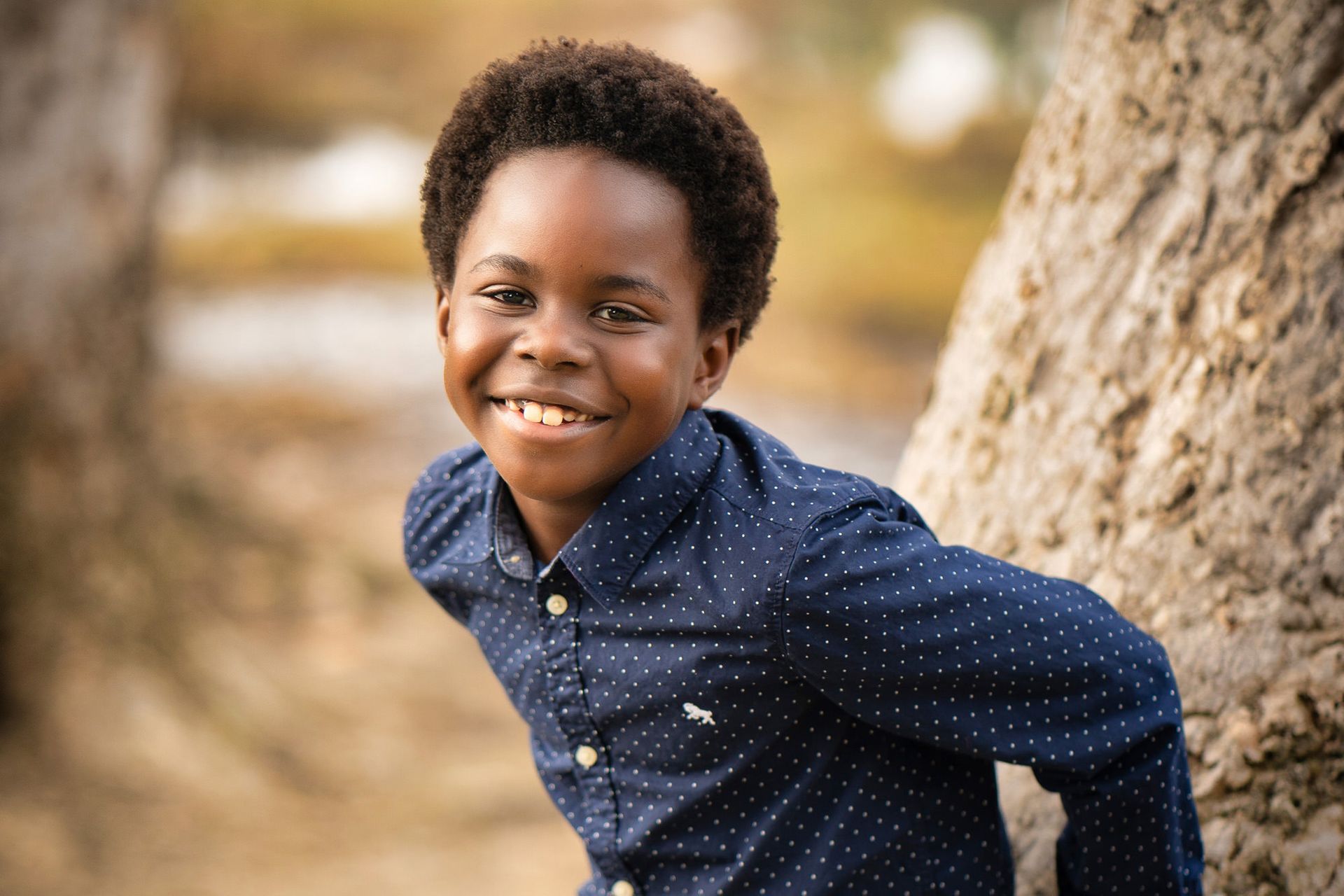 Orange County Fall Mini Sessions A young boy in a blue shirt is leaning against a tree and smiling in Huntington Beach Park.