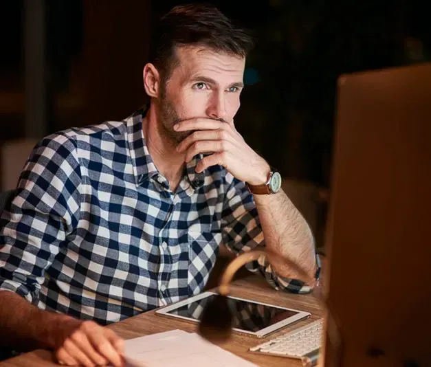 Man in plaid shirt thoughtfully looking at computer screen, hand to chin, at a desk with tablet and lamp.