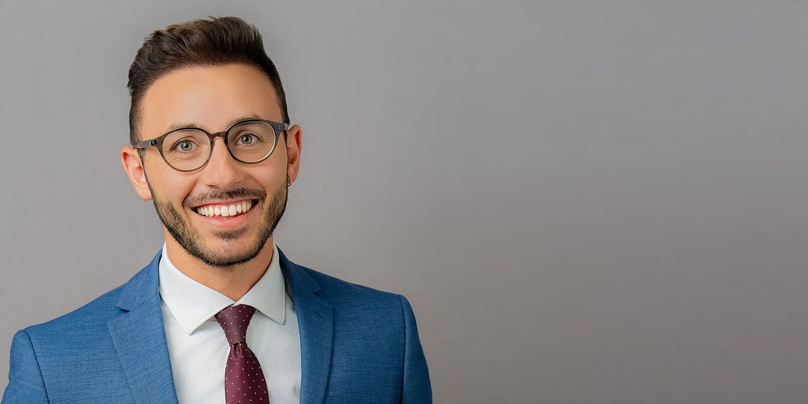 Man wearing glasses, smiling, in a blue suit and maroon tie against a gray background.