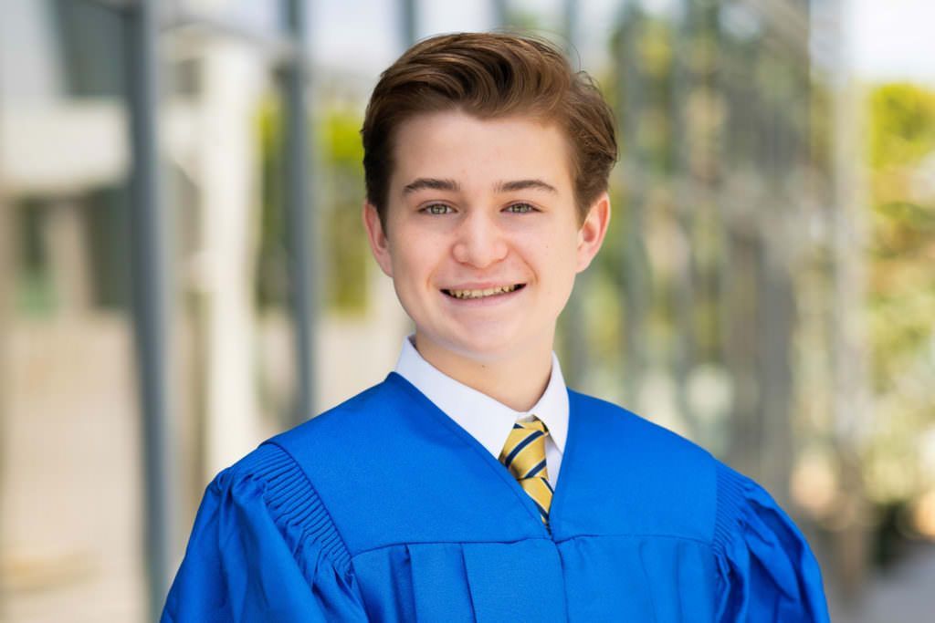 A young man in a blue graduation gown and tie is smiling for the camera at the Newport Beach Civic Center