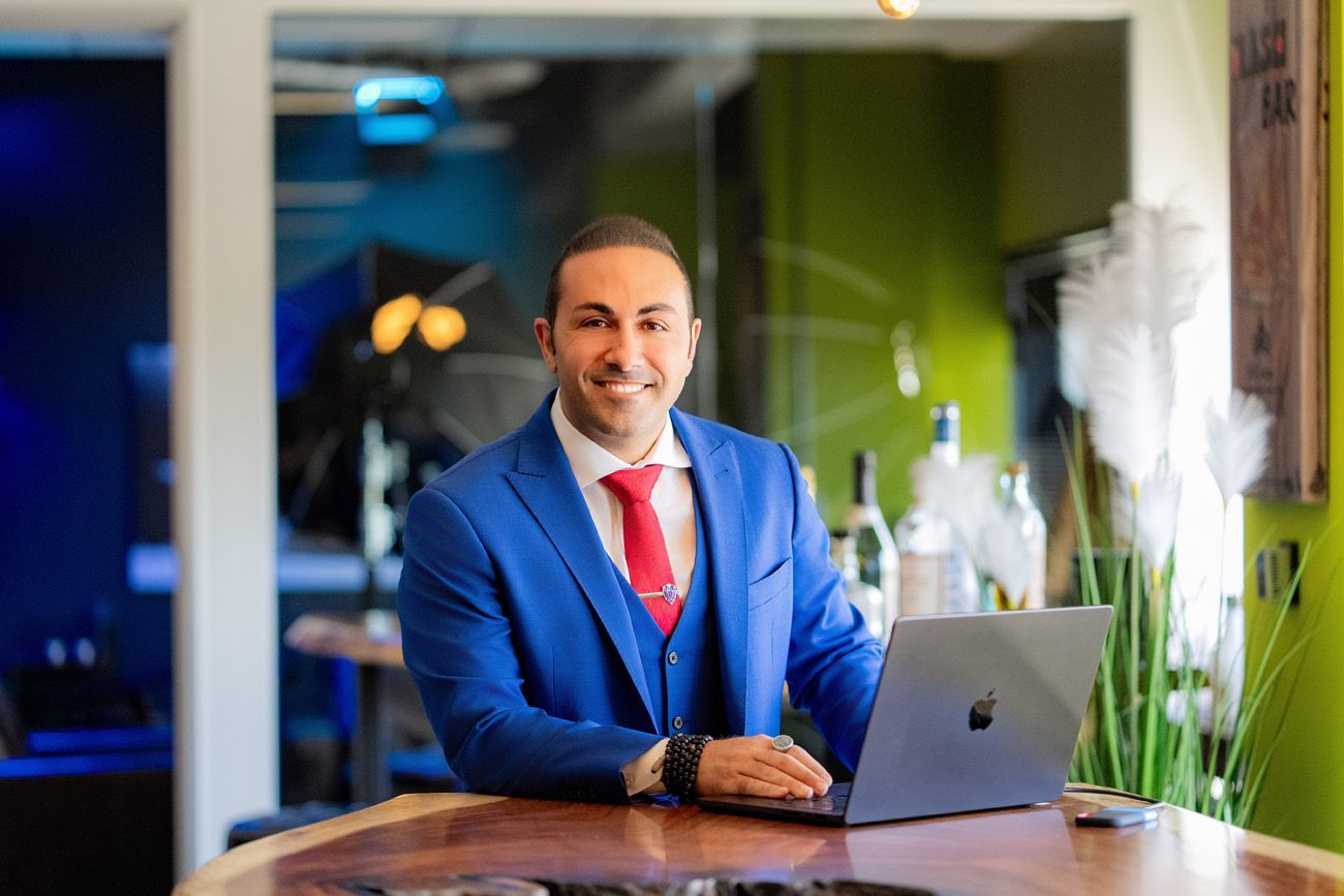 Man in blue suit, red tie, using laptop at a table, smiling. Modern office setting.