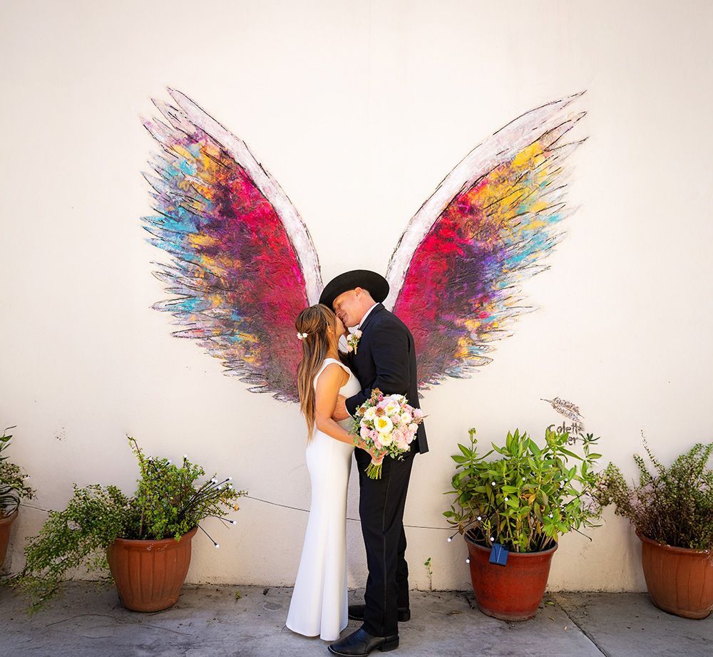 A bride and groom kiss in front of a wall with angel wings painted on it in Laguna Beach.