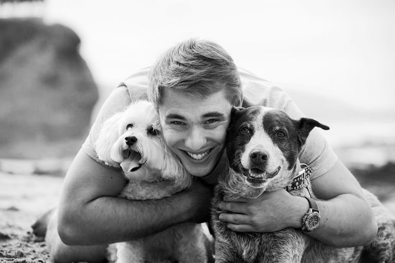 Shaws Cove Beach Portrait Ayoung man is hugging two dogs on the beach in a black and white photo.