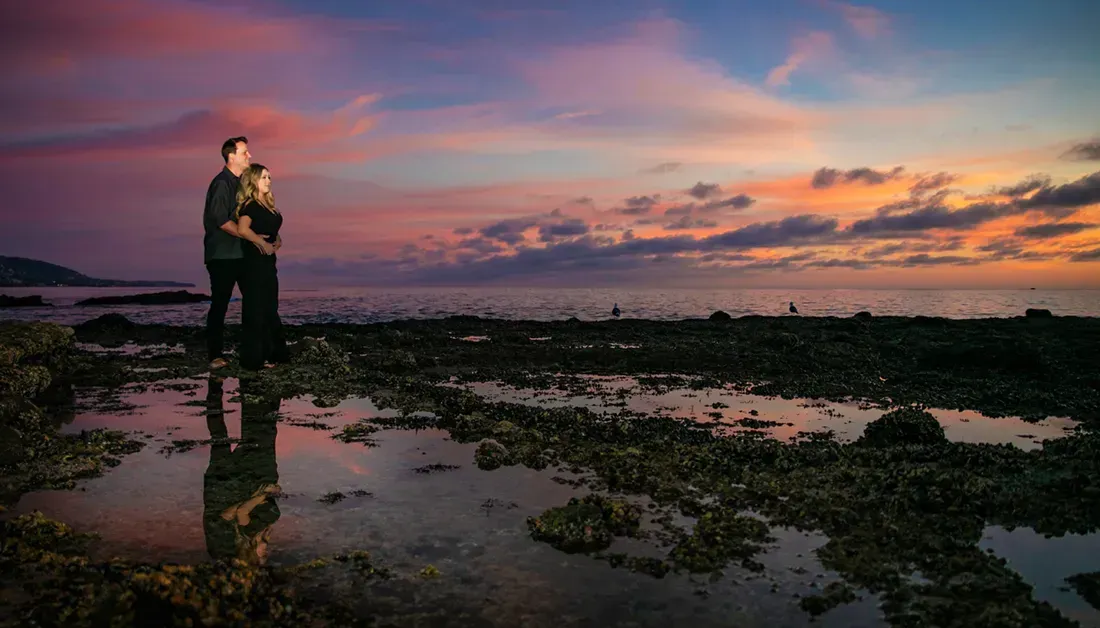 A man and a woman are standing on a beach at sunset in Laguna Beach.
