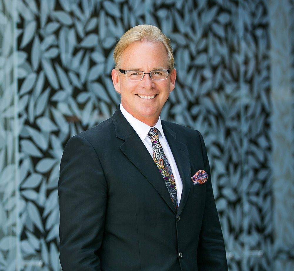 A man in a suit and tie is smiling in front of a wall with leaves on it