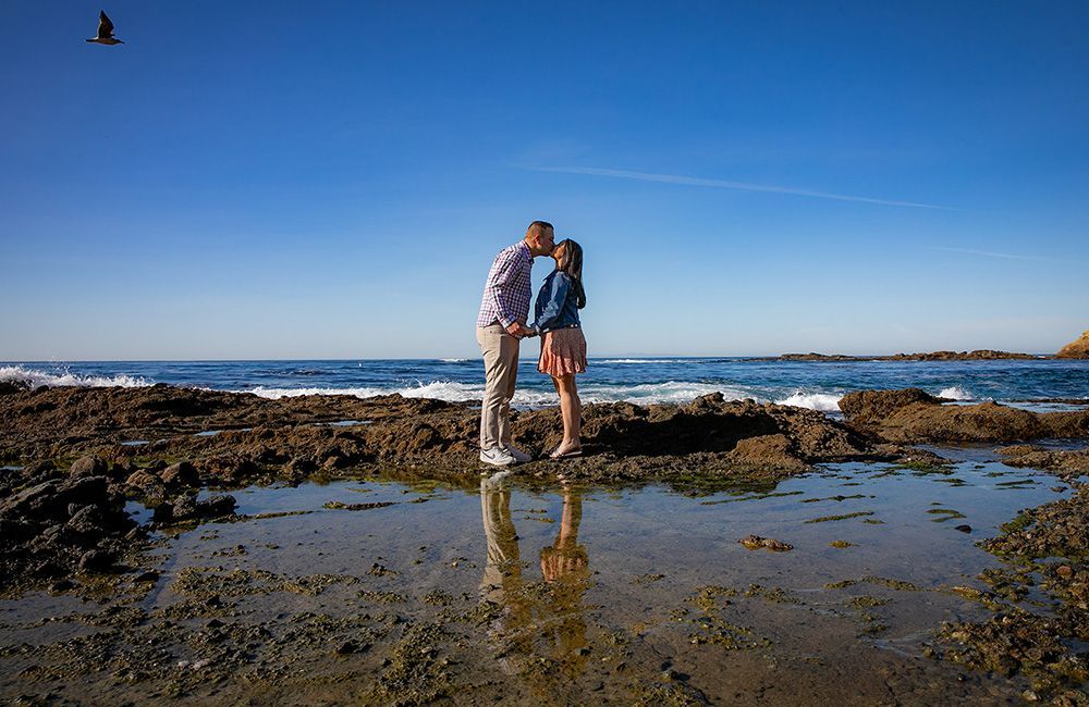 Engagement Photographer Dana Point A man and a woman are kissing on a rocky beach near the ocean.