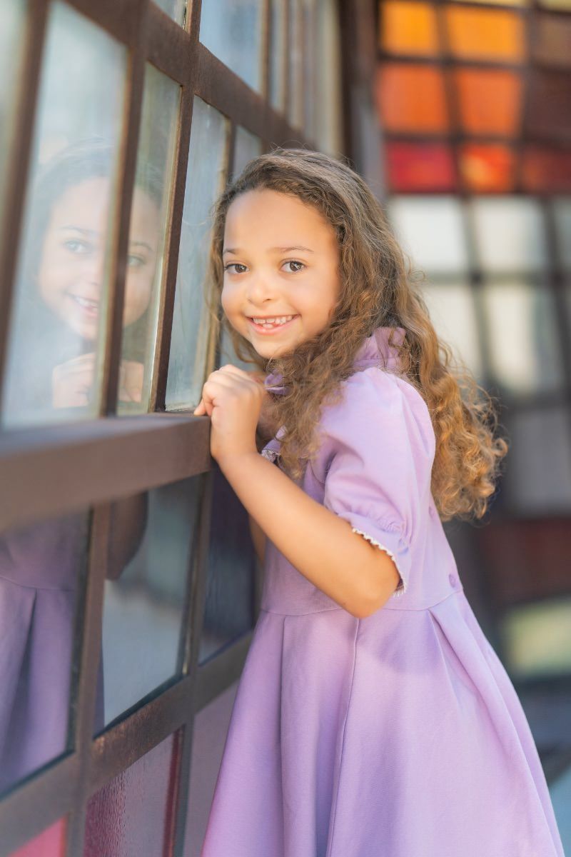 A little girl in a purple dress is standing next to a stained glass window in Noguchi Garden. 