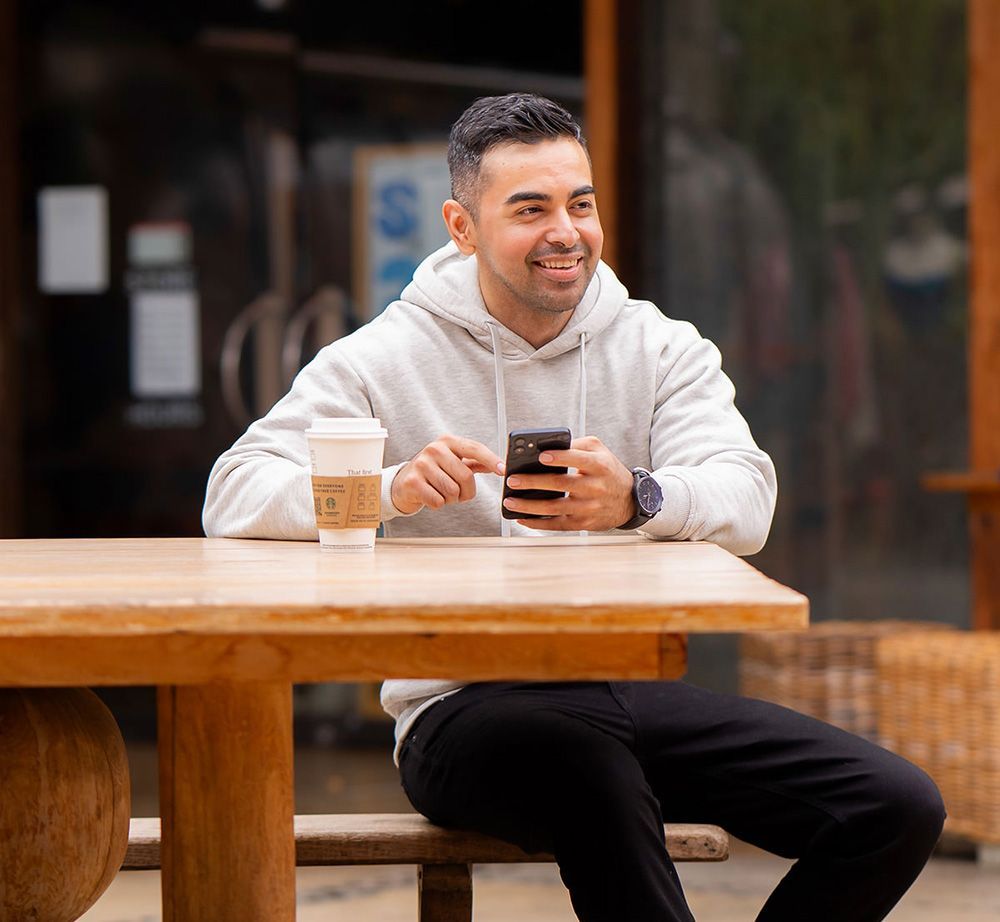 A man is sitting at a table with a cup of coffee and a cell phone for a Hinge Profile Photo