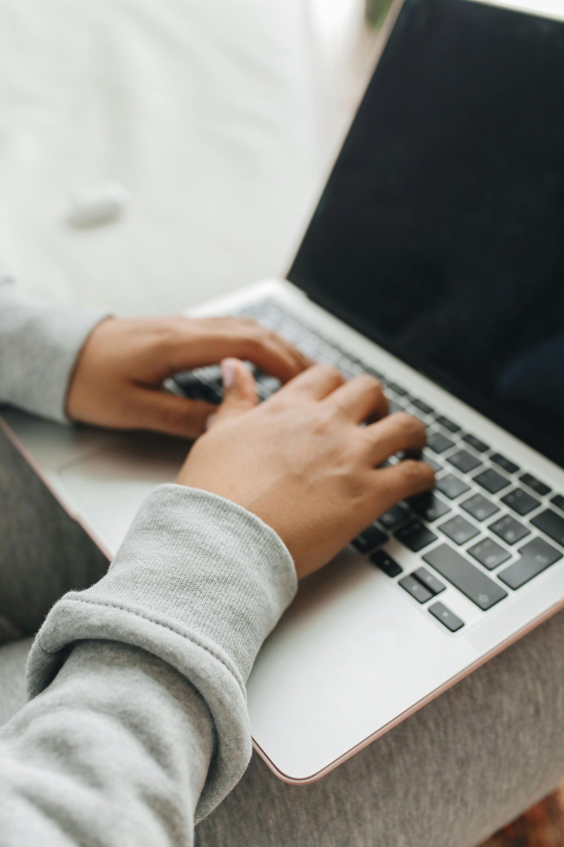 Hands typing on a silver laptop computer held on a person's lap, wearing a light gray long-sleeved shirt.