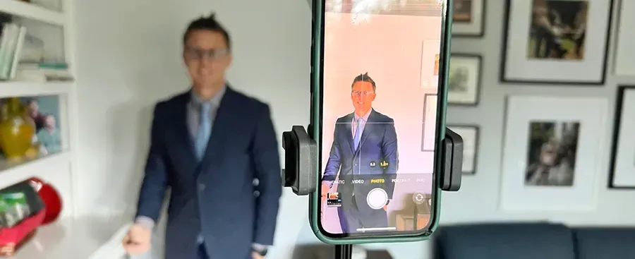 A man in a suit is standing in taking a selfie with a cell phone and remote control