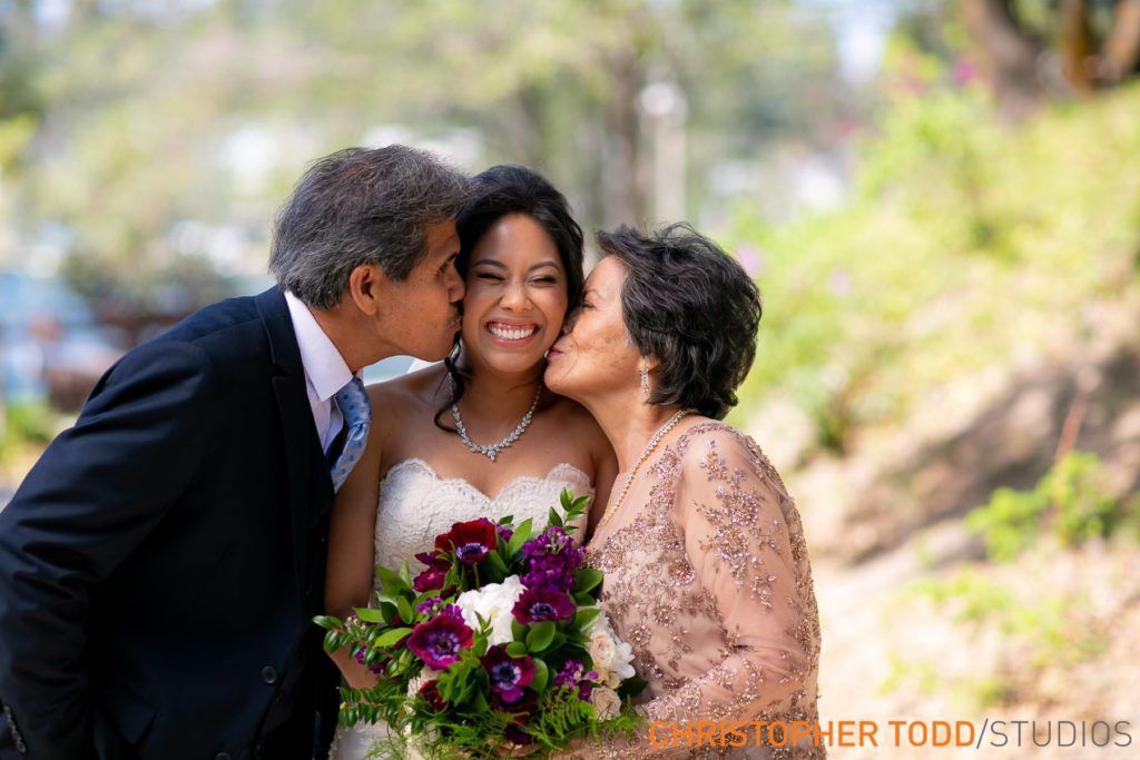 A bride and her parents are kissing her on the cheek.