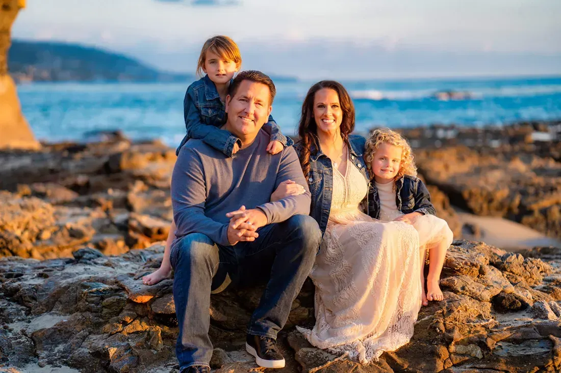 A family is posing for a picture on the beach with a dog and a wooden pier in background
