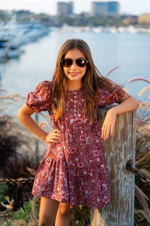 A young girl in a dress and sunglasses is leaning against a wooden fence.