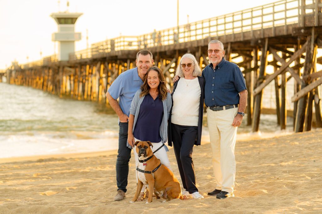 A family is posing for a picture on the beach with a dog and a wooden pier in background