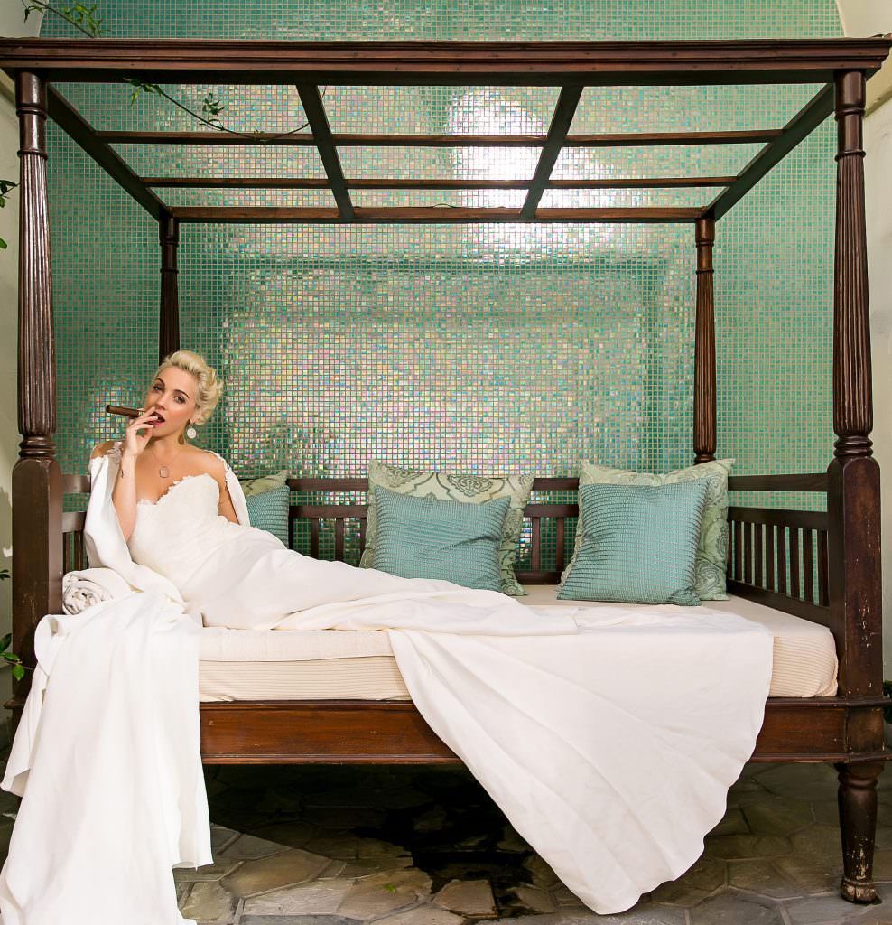 A Bride in a white dress is laying on a canopy bed smoking a cigar