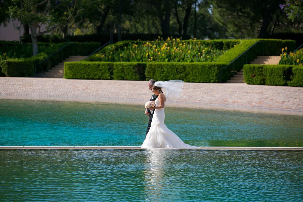 A bride and groom are walking across a pool of water at Soka University in Aliso Viejo.
