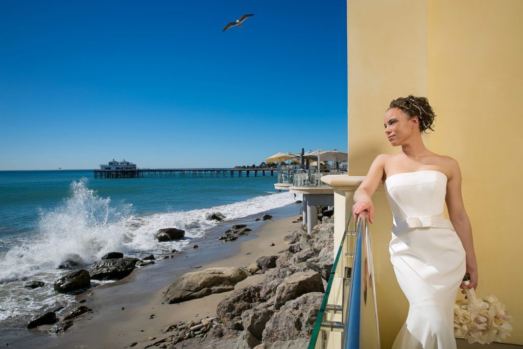 A bride in a white dress is standing on a balcony overlooking the ocean.