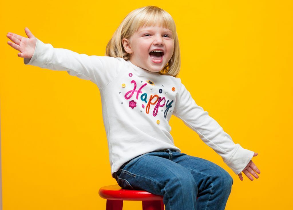 A little girl is sitting on a red stool with her arms outstretched.