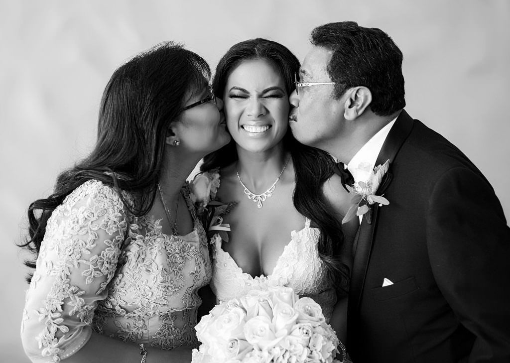 A black and white photo of a bride and her parents kissing her on the cheek.