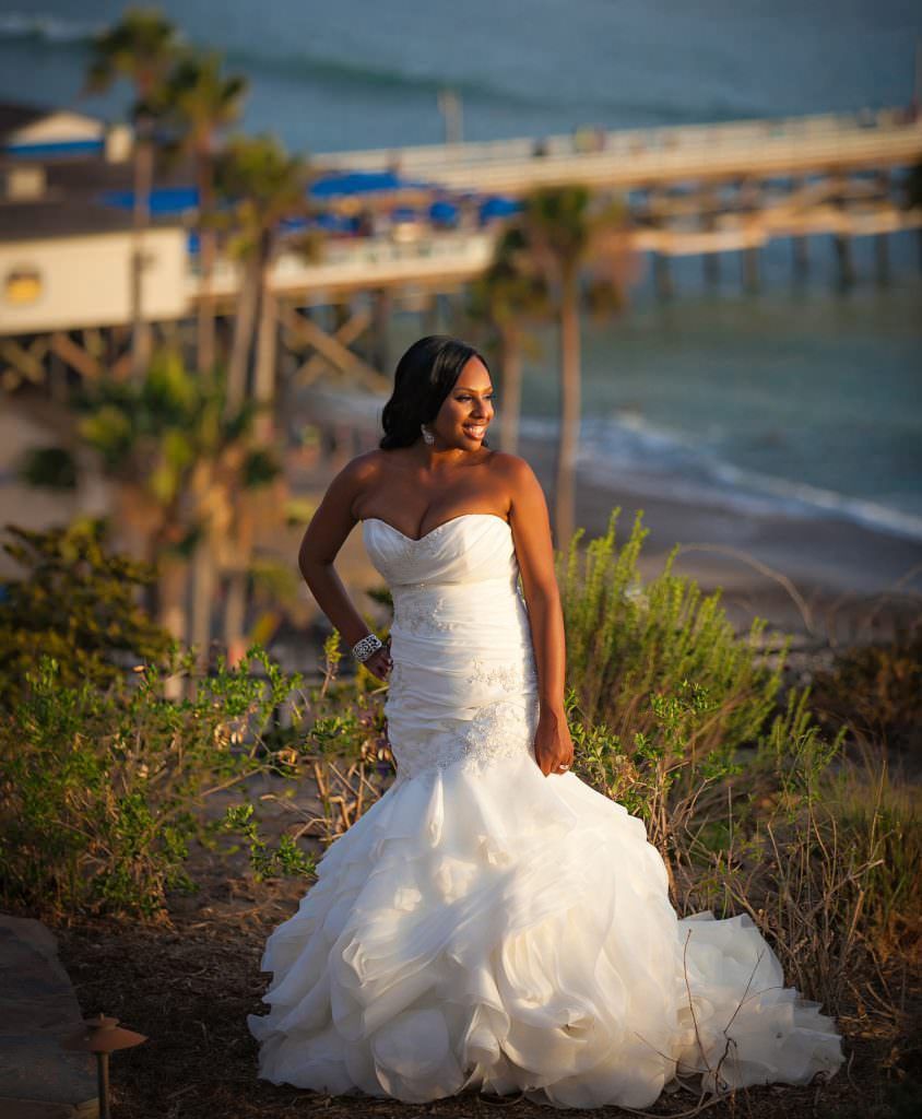 A San Clemente Bride in a wedding dress is standing on a hill near the ocean.