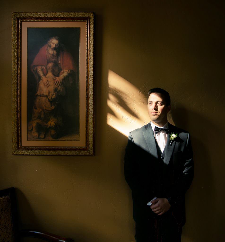 A Catholic groom  in a suit and bow tie is standing in front of a painting on a wall.