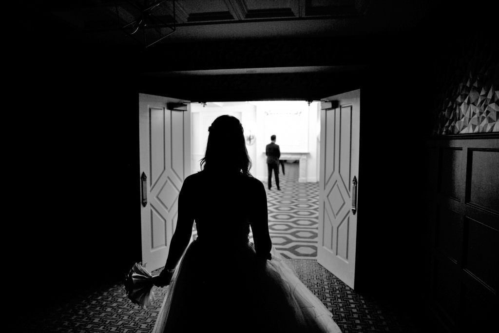 A bride and groom are walking down a hallway in a black and white photo for a first look before the ceremony