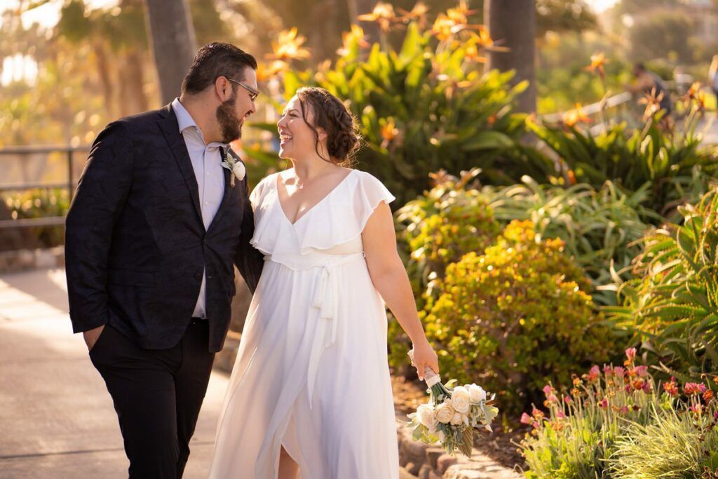 A bride and groom are walking down a sidewalk in a garden in Heisler Park Laguna Beach. 
