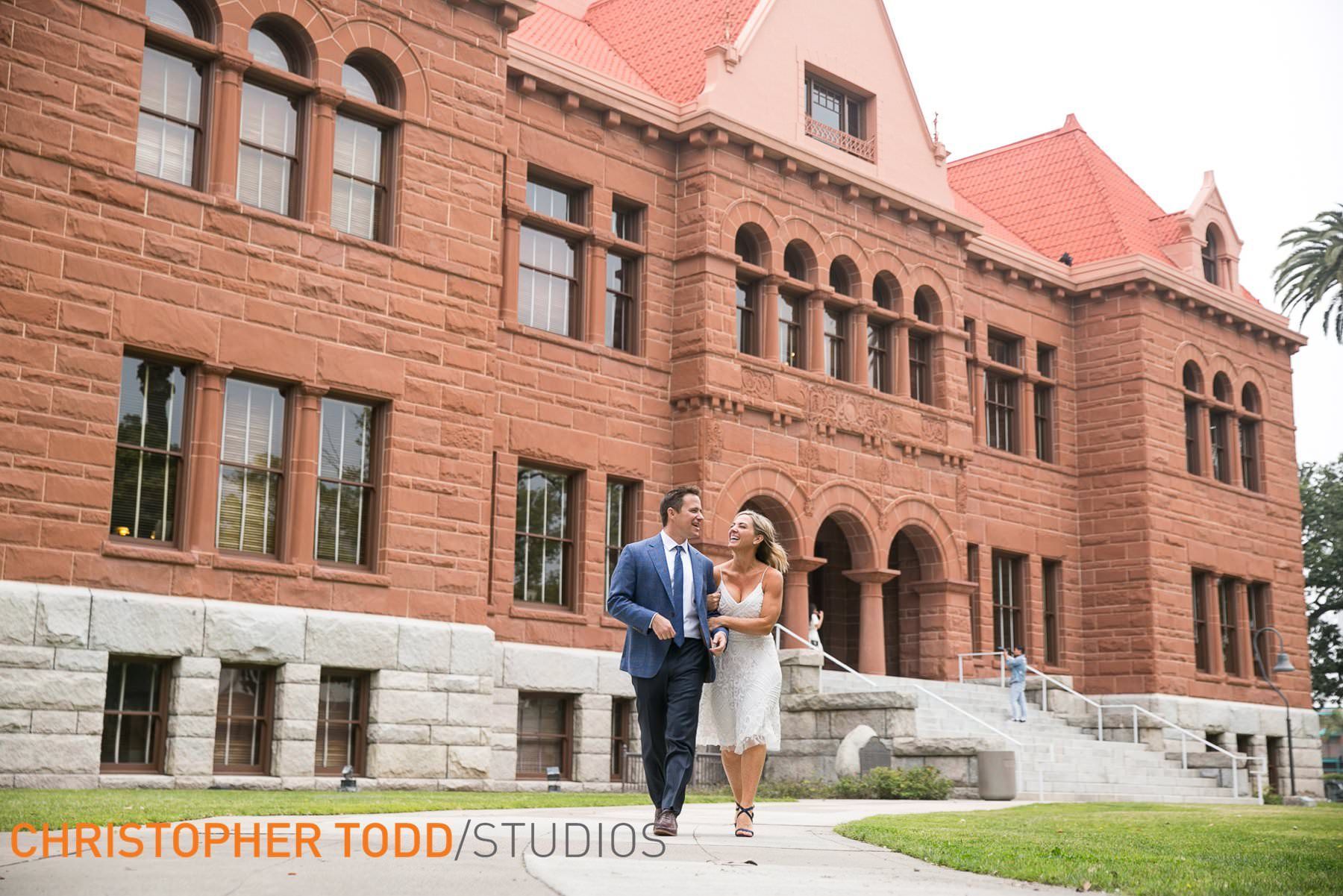 Old Orange County Courthouse Wedding A bride and groom are walking in front of a the Old Orange County Courthouse in Santa Analarge brick building.