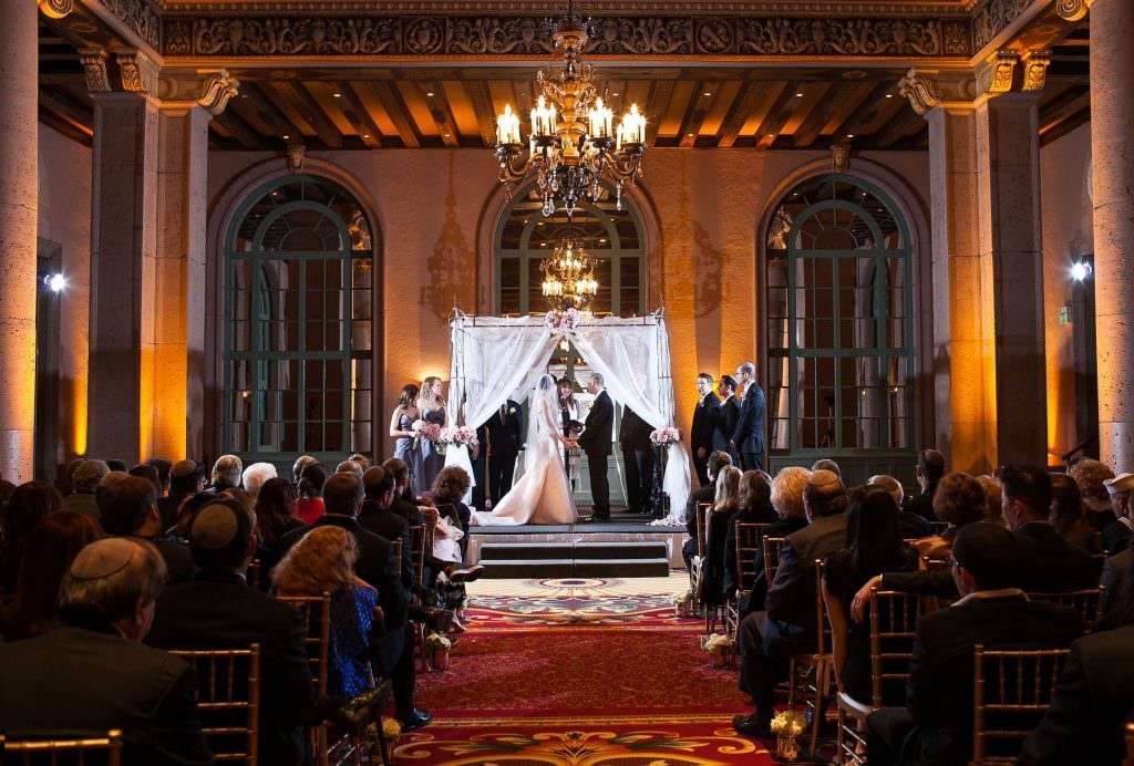 A Jewishbride and groom are getting married in front of a crowd of people at the Biltmore Hotel in Los Angeles