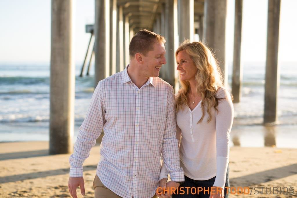 Engagement Photo Huntington Beach Pier A man and a woman are holding hands on the beach under the Huntington beach pier.