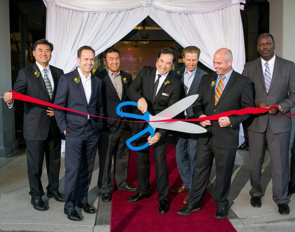 A group of men in suits are cutting a red ribbon with giant scissors