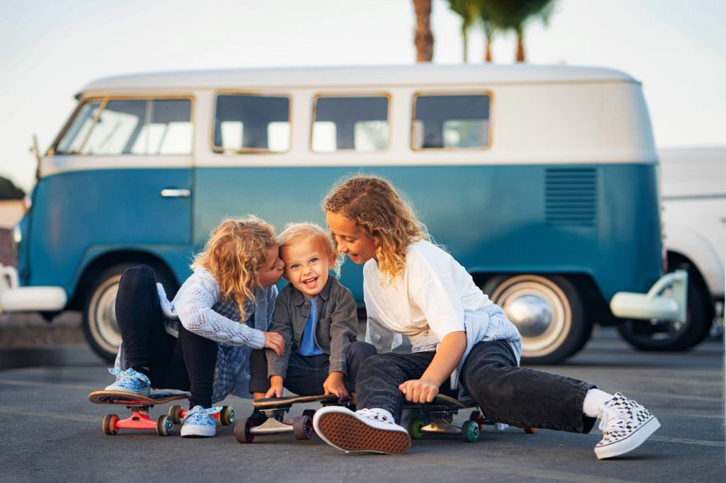 Three children are sitting on skateboards in front of a blue and white van.