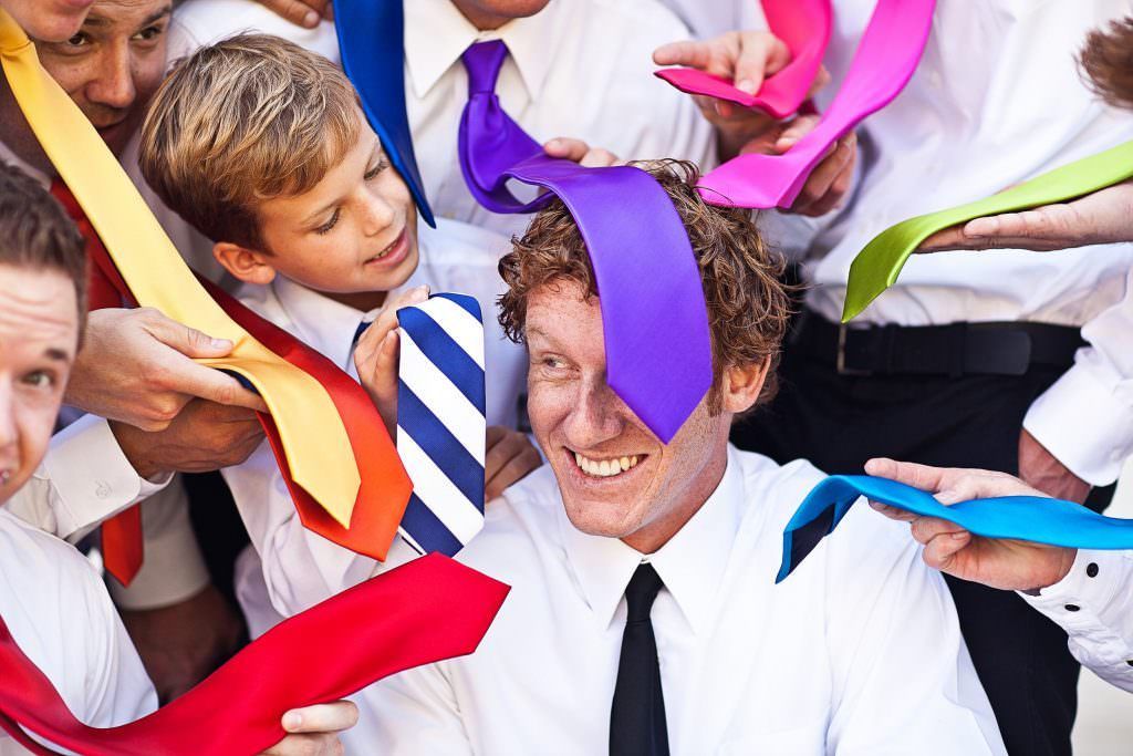 A groom with a purple tie on his head is surrounded by groomsmen holding colorful ties