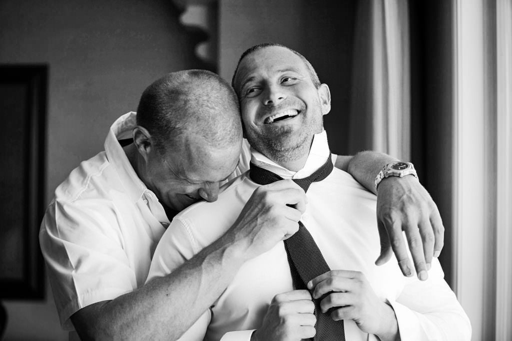 A man is helping a groom tie his tie befoe his wedding
