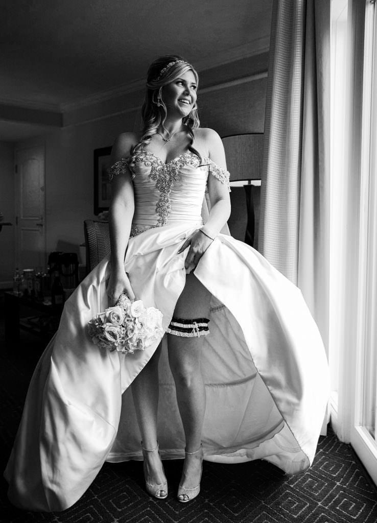 A black and white photo of a bride in a wedding dress standing in front of a window.