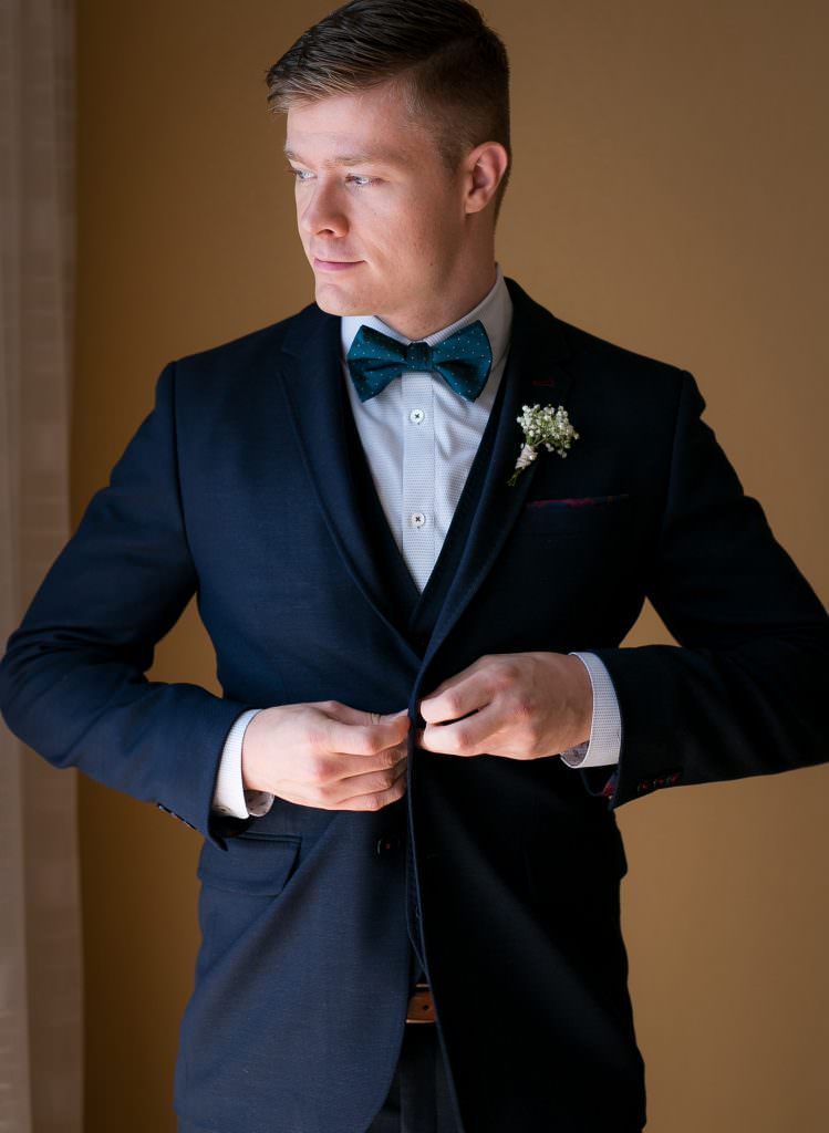 A  groom in a suit and bow tie is adjusting his jacket.