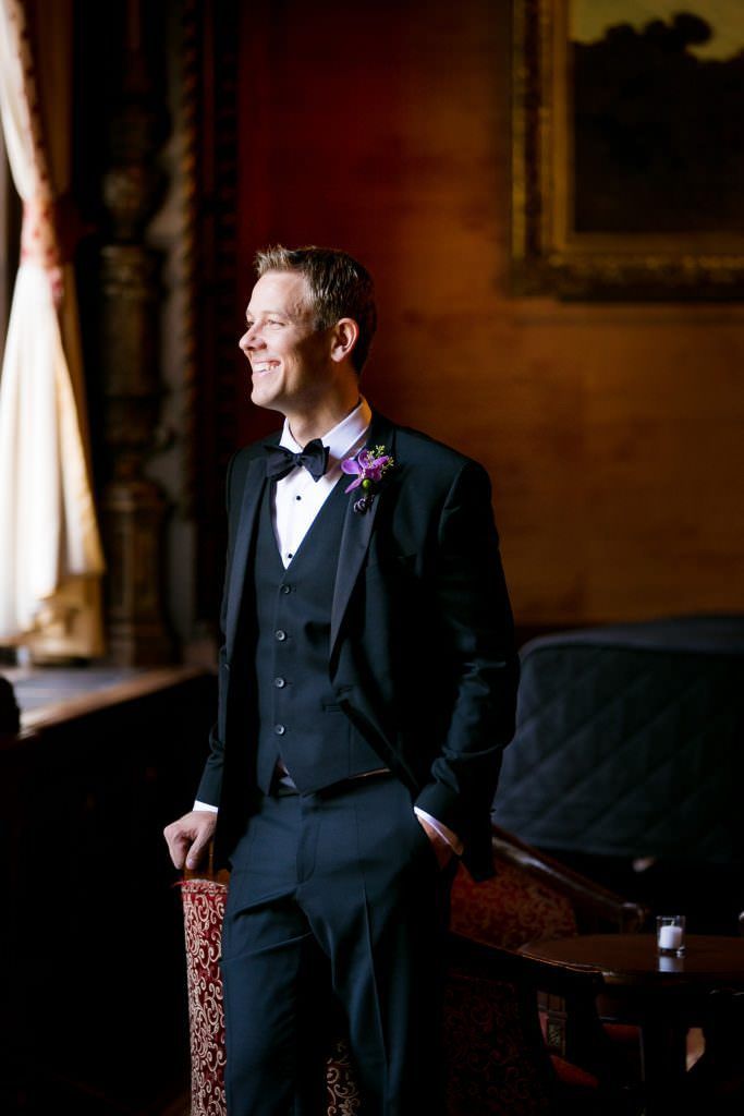A Groom in a tuxedo and bow tie is standing in front of a window.