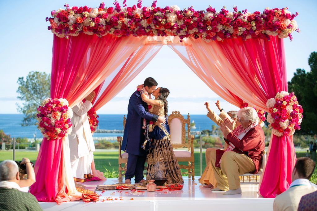 A bride and groom are kissing under a pink canopy at their Indian wedding ceremony.