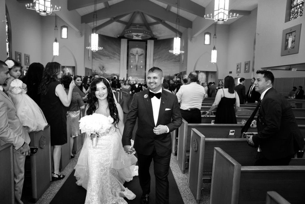 A Catholic bride and groom are walking down the aisle of a church holding hands.