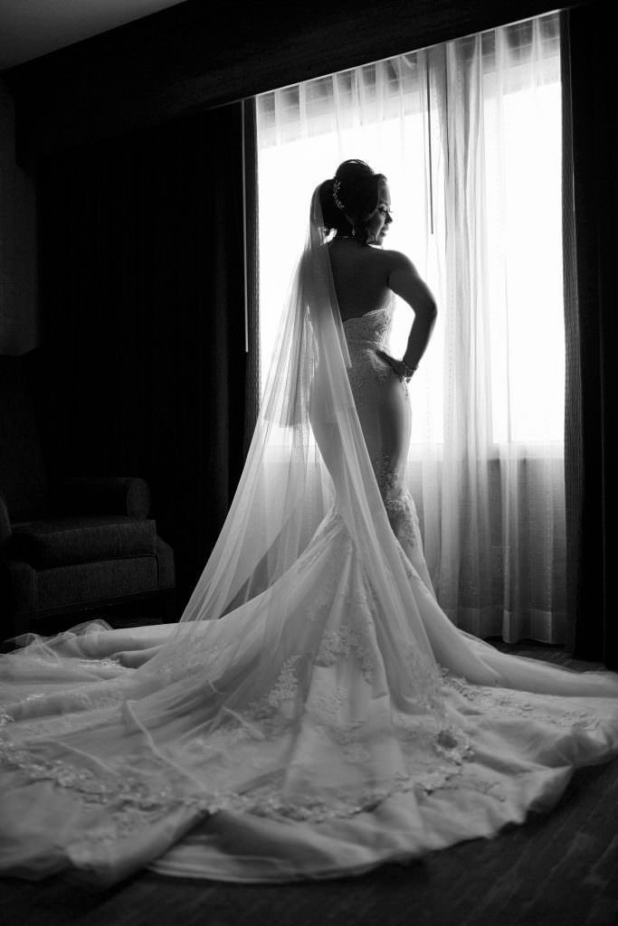 A black and white photo of a bride in a wedding dress standing in front of a window.