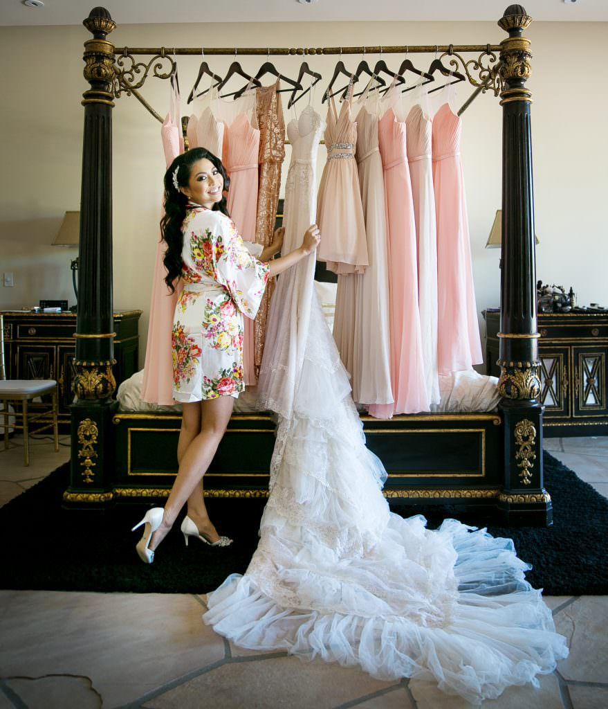 A woman in a robe is standing next to a bed holding a wedding dress.