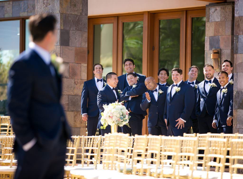 A groom in a suit is standing in front of a group of men in tuxedos.