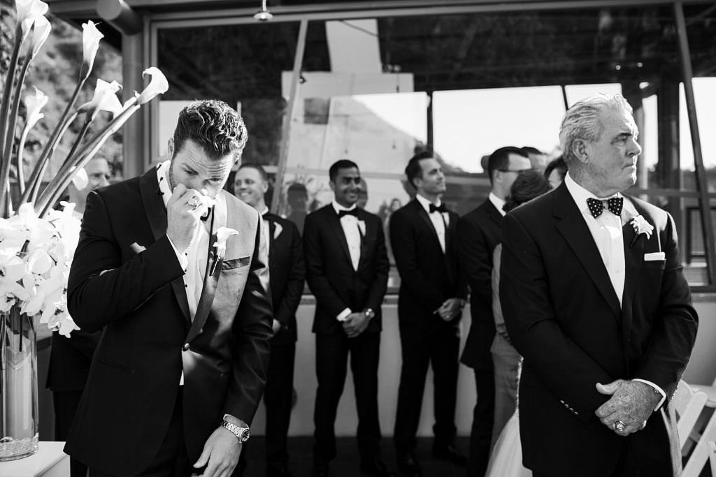 A groom, tears up while his bride walks down the aisle and her dad looks on