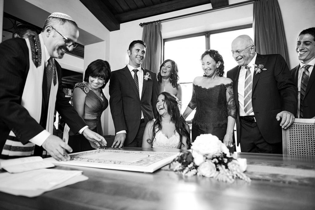 A black and white photo of a group of Jewish people standing around a table signing a Katuba .