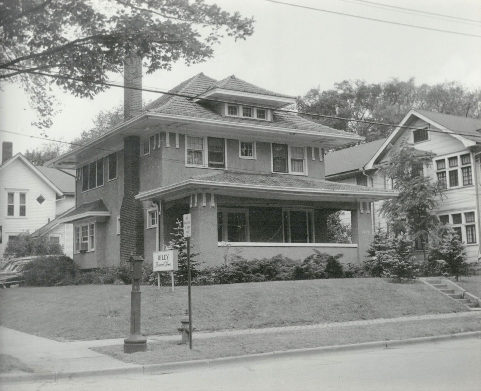 Sepia-toned photo of a two-story Prairie-style house with a wide front porch, a tiered roof, and a sign in the yard.