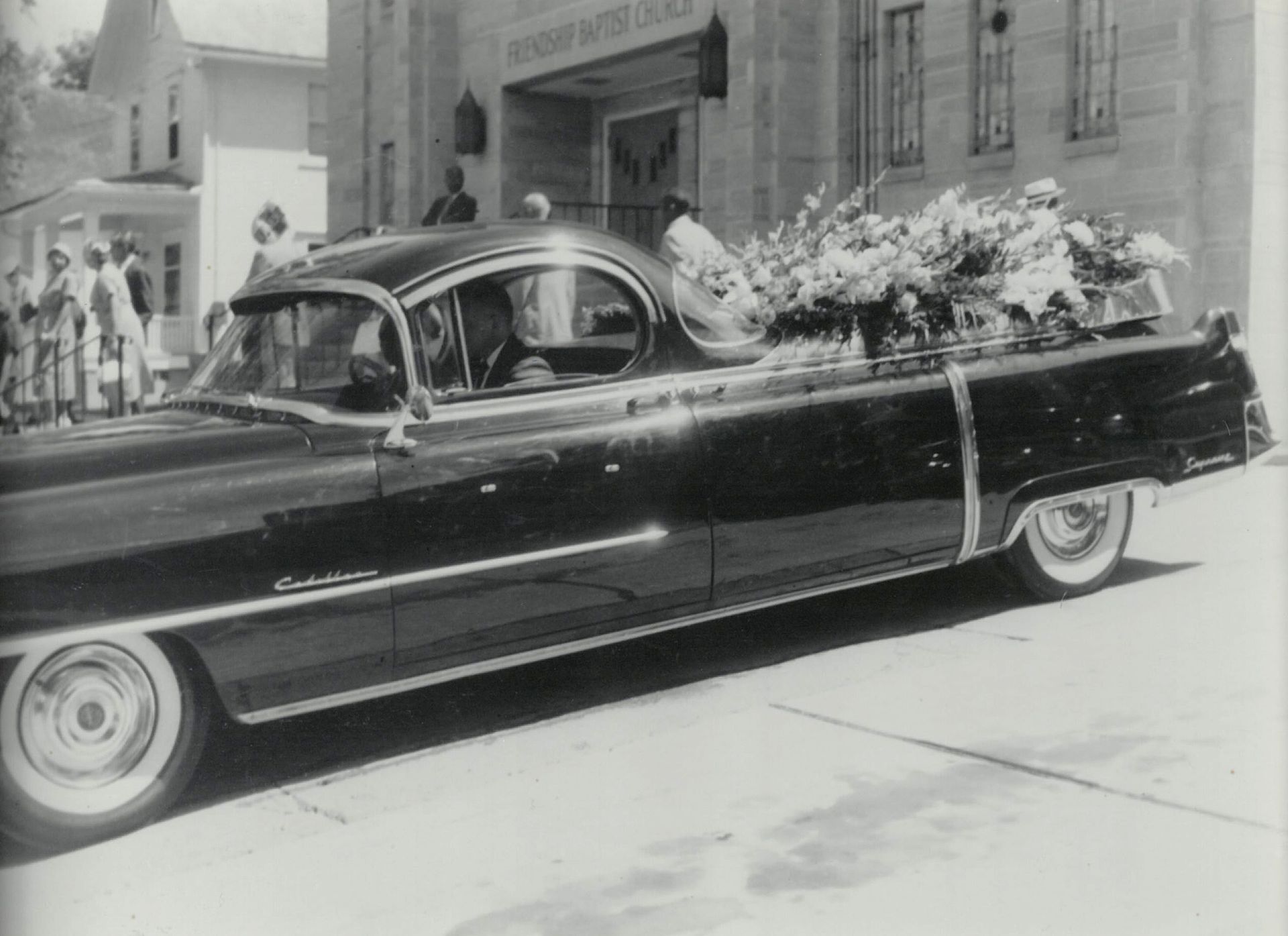 A vintage car carrying large floral arrangements drives past a stone building in a black-and-white framed photograph.
